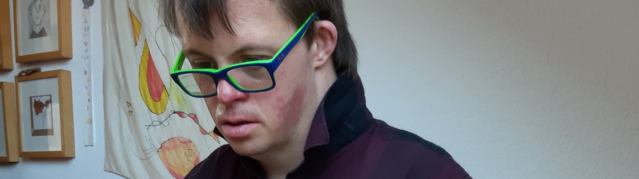 Ein Mann mit Down-Syndrom mit kurzen Haaren und Brille sitzt an einem Laptop und tippt.