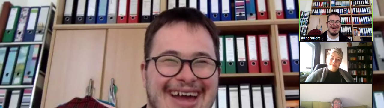Ein Mann mit Down-Syndrom mit Brille und Bart sitzt vor einem Bücherregal und lacht. Man sieht weitere Personen, die in einer Videokonferenz miteinander verbunden sind.