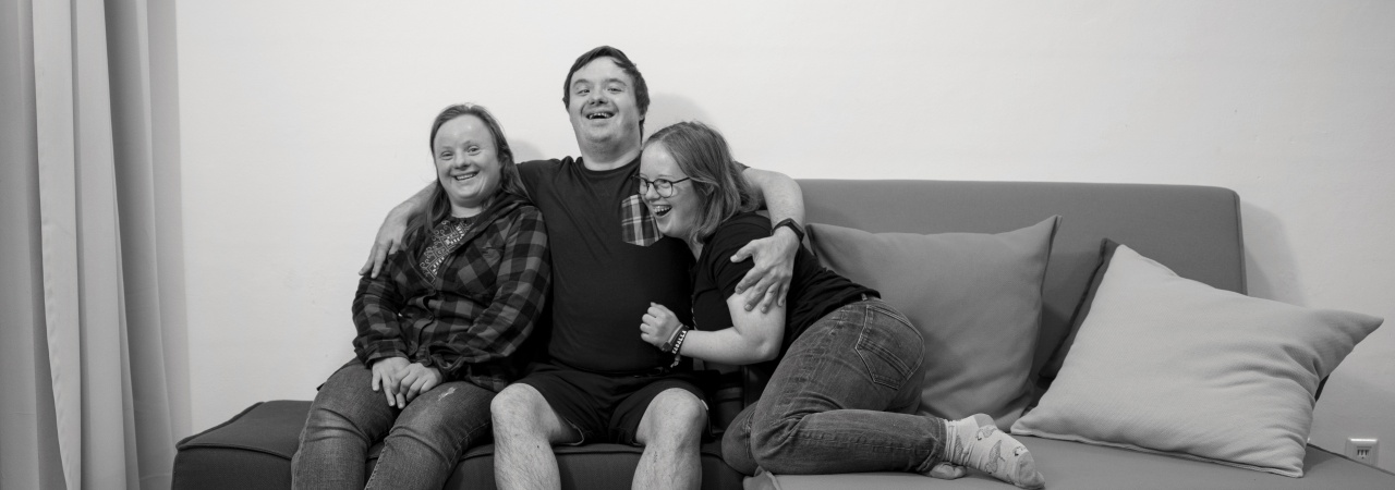 Schwarz-weiß-Foto von 3 Personen mit Down-Syndrom auf einer Couch. Ein Mann sitzt lächelnd in der Mitte und hält die beiden Frauen im Arm.