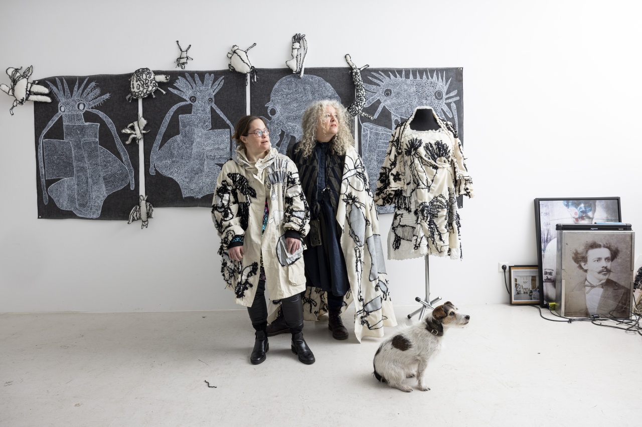 2 Frauen in kunstvoll bestickter Kleidung stehen nebeneinander in einem Atelier. Auf dem Boden neben ihnen sitzt ein Hund.