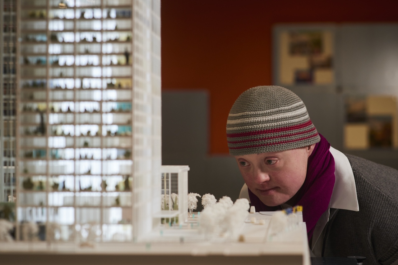 Eine Frau mit Down-Syndrom kniet vor einem Exponat der Ausstellung und betrachtet es konzentriert. Sie trägt eine gehäkelte Mütze.