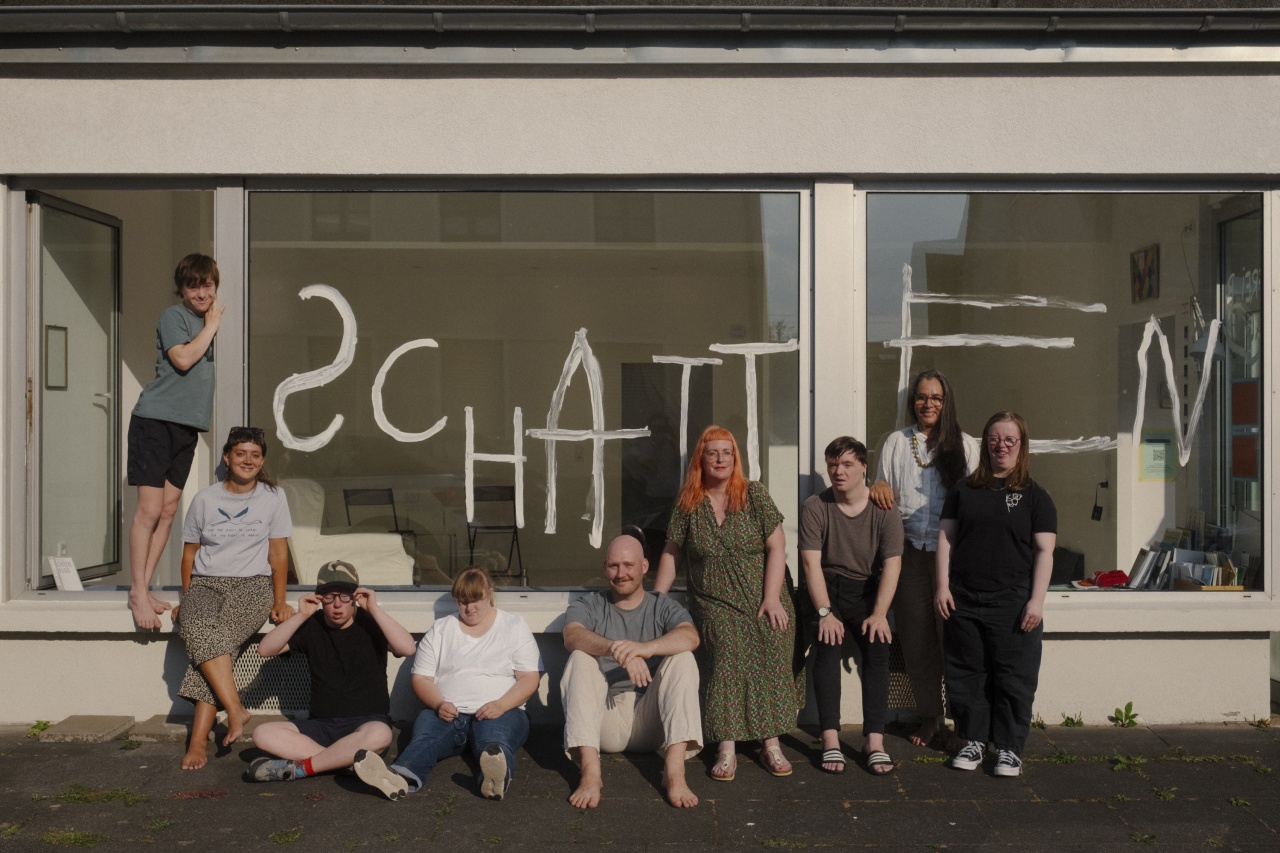 Eine Gruppe von Menschen mit und ohne Down-Syndrom steht im Freien vor einem Gebäude. Hinter ihnen eine Scheibe, an die mit weißer Farbe Schatten geschrieben ist.