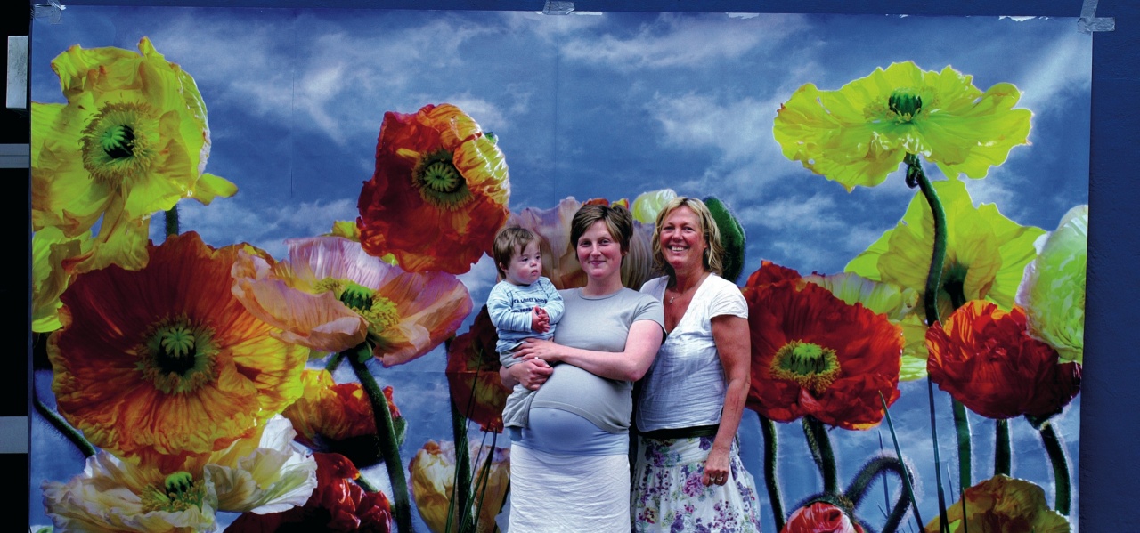Eine Schwangere mit Baby mit Down-Syndrom auf dem Arm und ihrer Mutter vor einer Blumentapete