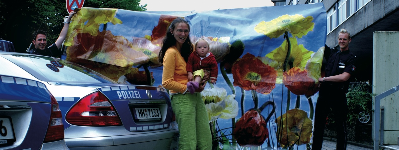 Eine Mutter mit ihrem Baby mit Down-Syndrom steht vor einer Blumentapete, die zwei Polizisten festhalten
