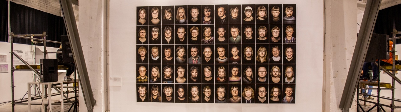 62 Portraitfotos von Menschen mit Down-Syndrom vor einer Wand in einem Ausstellungsraum