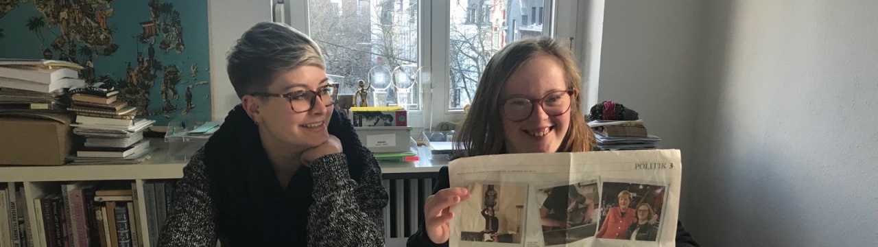 2 Frauen, eine mit und eine ohne Down-Syndrom, sitzen nebeneinander. Eine der beiden hält einen Zeitungsartikel in die Kamera.