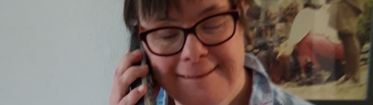 Eine Frau mit Down-Syndrom mit kurzen Haaren und Brille. Sie telefoniert und sitzt vor einem Gemälde.