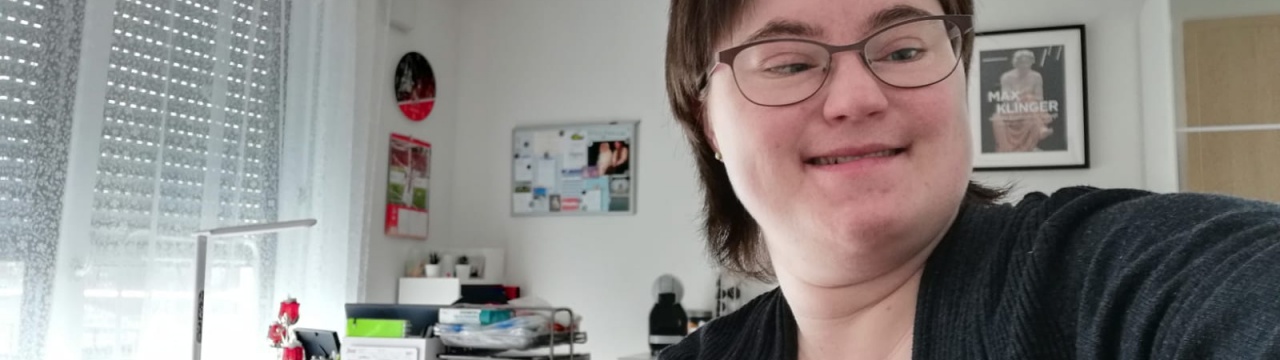 Ein Selfie einer Frau mit Down-Syndrom. Sie hat braune Haare und trägt eine Brille.