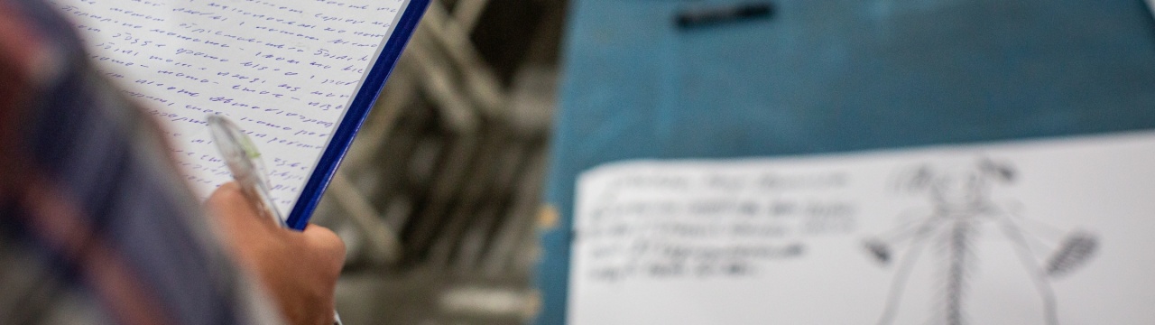 Eine Person in kariertem Oberteil hält eine Klemmmappe mit einem handgeschriebenen Text in kyrillischen Buchstaben in der Hand. Daneben liegt eine Zeichnung einer Figur, ebenfalls mit handgeschriebenem Text daneben.