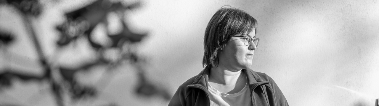 Schwarz-wieß-Foto einer Frau mit Down-Syndrom mit ernstem Blick. Sie steht in der Sonne und schaut zur Seite. In der Hand hält sie eine Schutzmaske.