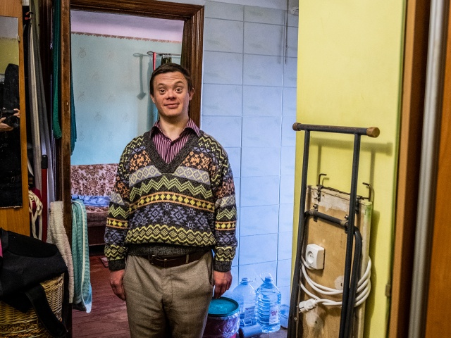 Valentin Radschenko in einem gemusterten Strickpullover, lächelnd in seiner Kiewer Wohnung. Neben ihn lehnt ein Bügelbrett an der Wand.