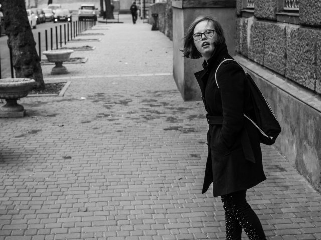 Schwarz-weiß-Foto einer Frau mit Down-Syndrom. Sie läuft in einem schwarzen Mantel durch die Straßen Kiews und dreht sich über die Schulter zur Kamera.