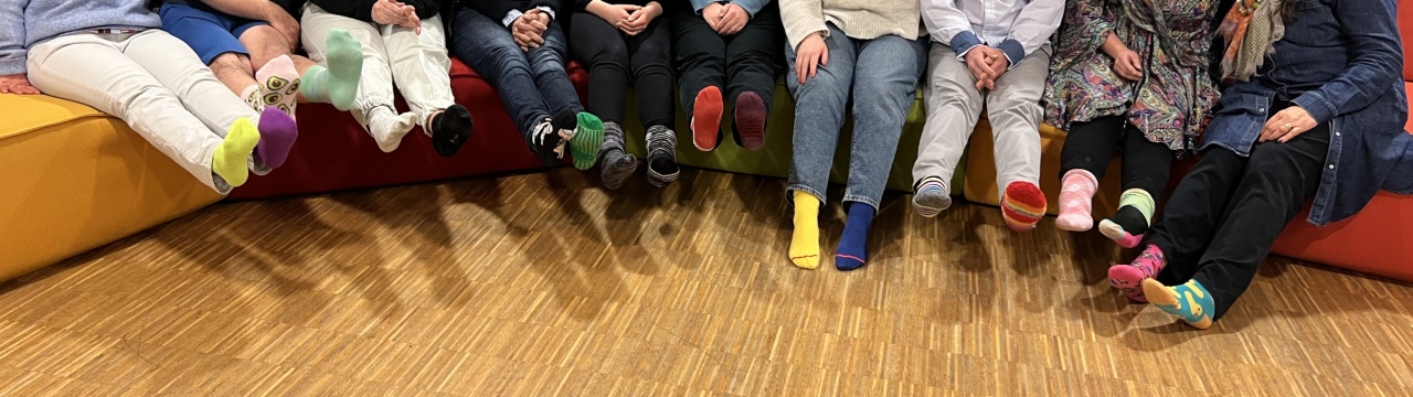 Viele Menschen, fast alle von ihnen mit Down-Syndrom, sitzen auf bunten Sofas und tragen zwei verschiedene Socken an den Füßen.