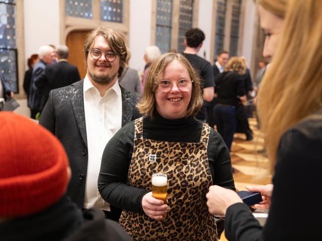 Natalie Dedreux hält ein Kölsch in der Hand. Hinter ihr steht ein Mann mit Down-Syndrom im Anzug.