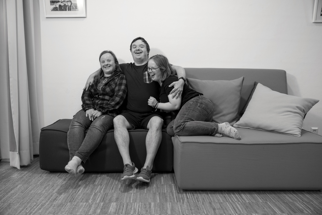 Zwei Frauen und ein Mann mit Down-Syndrom sitzen zusammen auf der Couch und lachen. Der Mann sitzt in der Mitte und hat seine Arme um die beiden Frauen gelegt.