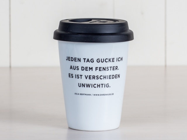 Kaffeebecher aus weißem Porzellan mit schwarzem Silikondeckel, auf der Frontseite steht das Zitat