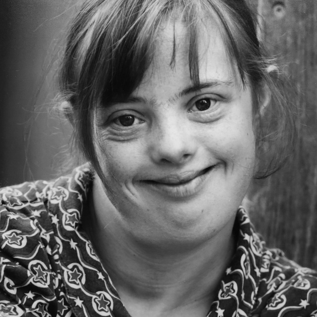Eine junge Frau mit Down-Syndrom mit fransigem Pony. Sie lächelt breit in die Kamera.