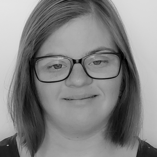 Schwarz-weiß-Foto einer jungen Frau mit Down-Syndrom mit glatten, schulterlangen Haaren und Brille.